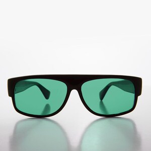 May include: Black sunglasses with rectangular frames and green lenses. The sunglasses have a thick, solid frame design. The lenses are a vibrant green color, offering a bold contrast against the black frame. The sunglasses are displayed on a reflective surface.
