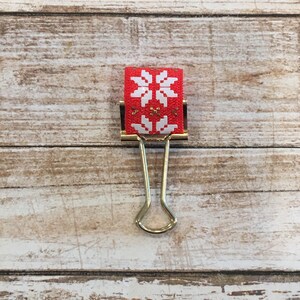 May include: A gold binder clip with a red ribbon with a white snowflake pattern.