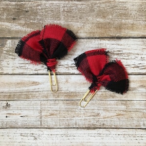 May include: Two gold paper clips with red and black buffalo plaid fabric bows attached. The bows are frayed at the edges.
