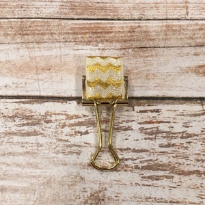 May include: A gold binder clip with a white and gold chevron patterned ribbon attached.