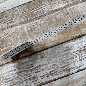 Puede incluir: Un rollo de cinta washi blanca con un patrón cuadrado negro. La cinta está sobre una superficie de madera.