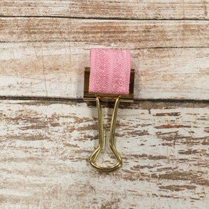May include: A gold binder clip with a pink elastic band attached to the top.