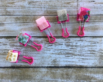 May include: Five decorative binder clips with pink handles and silver metal clips. The tops of the clips are covered in various patterned fabrics, including pink, floral, and glittery silver. The clips are arranged on a weathered, light brown wooden surface.