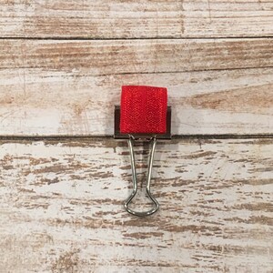May include: A silver binder clip with a red ribbon attached to it.