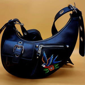 May include: Black crescent-shaped handbag with a shoulder strap and a small front pocket. The bag features a silver zipper and buckle. A colorful tattoo-style swallow and flower design is painted on the front.