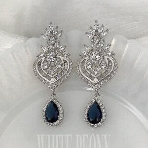 May include: A pair of silver-toned drop earrings with a heart-shaped design, encrusted with clear crystals. Each earring features a dark blue teardrop-shaped stone. The text "WHITE PEONY" is visible in the image.