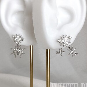 May include: A pair of silver starburst earrings with clear crystal accents. The earrings are displayed on a gold stand against a white background. The text "WHITE PEONY" is visible in the lower right corner of the image.