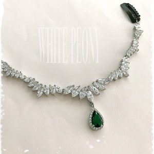 May include: A silver necklace with a teardrop-shaped emerald green gemstone pendant surrounded by smaller clear stones. The necklace is made of a chain of small clear stones arranged in a leaf pattern.