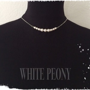 May include: A silver chain necklace with a row of white pearl beads. The necklace is displayed on a black mannequin bust. The text "WHITE PEONY" is printed below the necklace.
