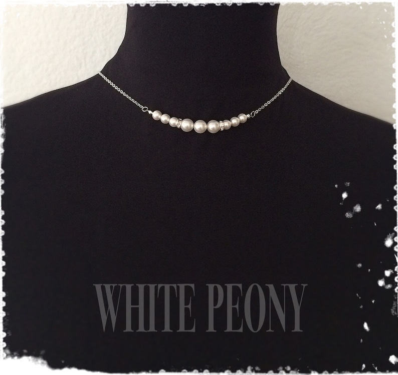 May include: A silver chain necklace with a row of white pearl beads. The necklace is displayed on a black mannequin bust. The text "WHITE PEONY" is printed below the necklace.