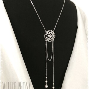 May include: A silver necklace with a delicate, ornate pendant featuring a floral design with clear crystals. The pendant hangs from a chain with two strands of pearls at the bottom.