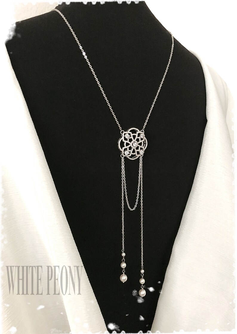 May include: A silver necklace with a delicate, ornate pendant featuring a floral design with clear crystals. The pendant hangs from a chain with two strands of pearls at the bottom.