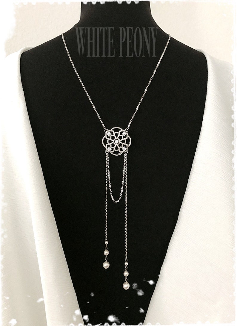 May include: A silver necklace with a delicate, ornate pendant featuring a circular design with small stones. The pendant is attached to two chains that end in pearl drops.