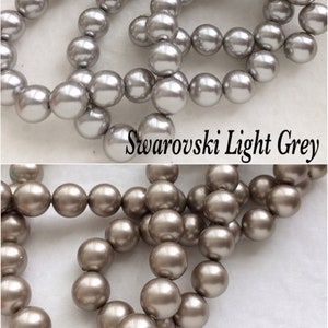 May include: Two strands of round, pearl-like beads. The top strand is light gray and labeled "Swarovski Light Grey". The bottom strand is a darker, silvery gray and labeled "Swarovski Platinum".