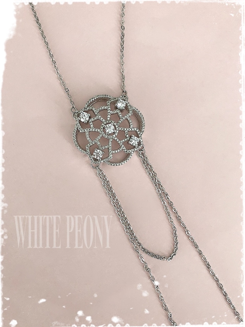 May include: A silver necklace with a delicate, circular pendant featuring a geometric design with small clear stones. The pendant hangs from two chains.