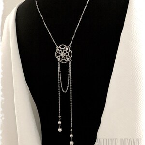 May include: A silver back necklace with a delicate, ornate pendant featuring a circular design with small clear stones. The pendant is attached to two chains that end in pearl-like beads.