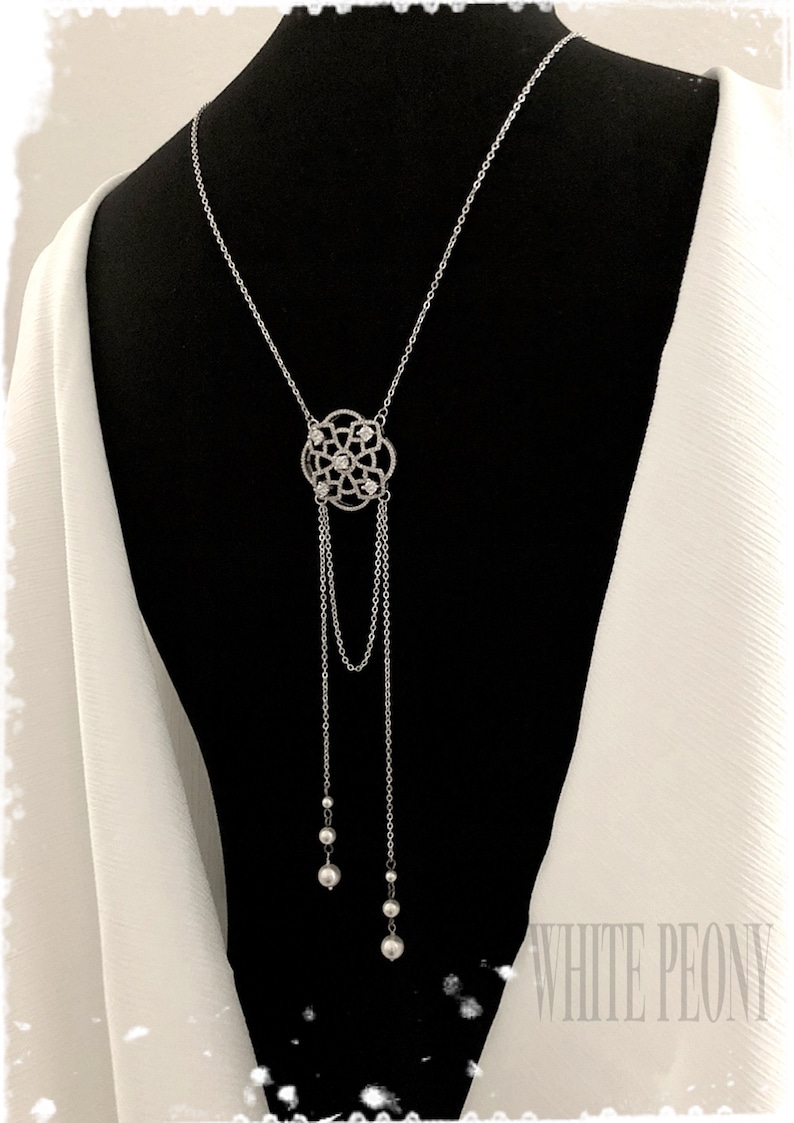May include: A silver back necklace with a delicate, ornate pendant featuring a circular design with small clear stones. The pendant is attached to two chains that end in pearl-like beads.