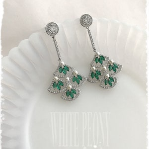 May include: A pair of silver dangle earrings with green and white stones. The earrings feature a fan-shaped design with multiple layers of stones.