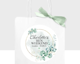 Op de afbeelding: Een witte papieren cadeauzak met een saliegroen lint aan het handvat. Een ronde sticker op de voorzijde toont een gouden ring, aquarel eucalyptusbladeren en de tekst Charlotte's HEN WEEKEND London ~ 03.09.21 met een klein groen hartje.