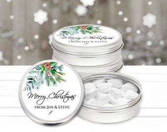 May include: A stack of round silver metal tins with lids featuring a watercolor illustration of pine branches and red berries. The text reads Merry Christmas FROM JEN & STEVE. One open tin reveals small white heart-shaped candies inside.