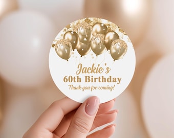 May include: A round, white sticker with gold text and gold balloon graphics. The text reads "Jackie's 60th Birthday Thank you for coming!" The sticker is held by a person with a blurred background of white and gold balloons.