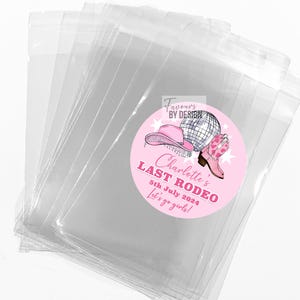 May include: A stack of clear cellophane bags with a pink circular sticker. The sticker features a pink cowboy hat, a disco ball, and a pink cowboy boot. Text on the sticker reads "Charlotte's Last Rodeo 5th July 2024 Let's go girls!"