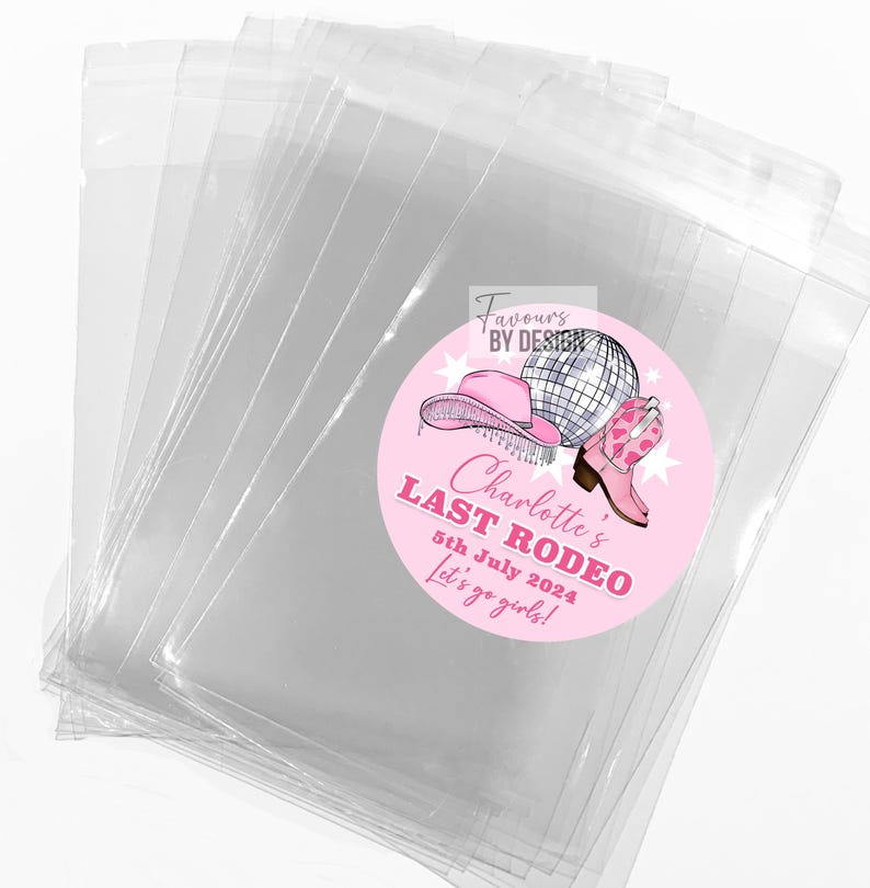 May include: A stack of clear cellophane bags with a pink circular sticker. The sticker features a pink cowboy hat, a disco ball, and a pink cowboy boot. Text on the sticker reads "Charlotte's Last Rodeo 5th July 2024 Let's go girls!"