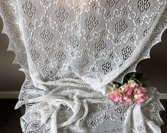 May include: A white crocheted shawl with a delicate lace pattern. The shawl is draped over a table with a bouquet of pink roses.