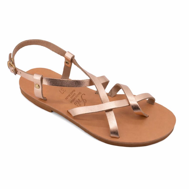 May include: Rose gold leather sandals with multiple straps and an ankle strap. The sandals have a flat sole and a buckle closure. The footbed is a light brown color. The sandals are size 37.