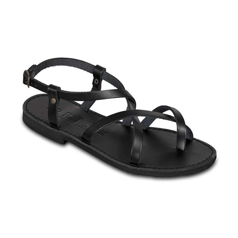 May include: Black leather sandals with multiple straps and a buckle closure. The sandals have a flat sole and a toe loop. The word "PRIS" is visible on the side of the sole. The sandals are a size 38.