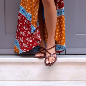 May include: Brown leather sandals with crisscross straps and ankle straps. The sandals are worn with a long, colorful patterned skirt in shades of red, orange, and blue. The sandals are on a white step.