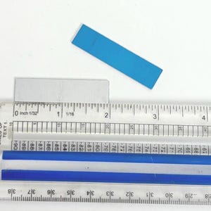 May include: A clear plastic ruler with inch and fractional markings, measuring a small, light blue rectangular object. The object is approximately 2 inches long. A smaller, gray rectangular object is also present.
