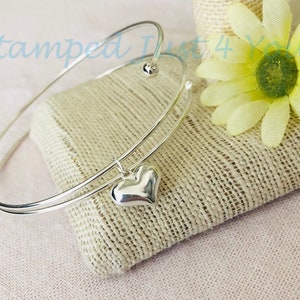 May include: A silver bangle bracelet with a charm in the shape of a heart. The bracelet is double-layered and has a small bead at the top.