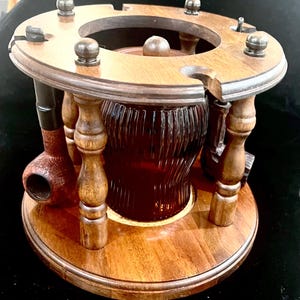 May include: A wooden pipe stand with a dark amber glass jar in the center. The stand has a circular base and a top with holes for pipes and accessories. The stand is made of polished wood and has a classic design.