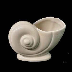 May include: A glossy, off-white ceramic planter shaped like a nautilus shell. The vessel features a smooth, curved exterior with a prominent spiral design on the side and a wide, open top for holding items, resting on a stable, oval-shaped base.