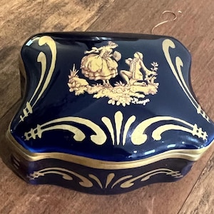May include: A navy blue ceramic trinket box with gold accents. The lid features a gold relief of two figures in a garden scene. Gold scrollwork decorates the edges of the box, which has a curved shape.