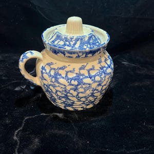 May include: A small, round, ceramic jar with a lid. The jar is cream-colored with a blue, spatter-like pattern. It has a handle and a lid with a small knob. The jar is likely for storing food or other small items.