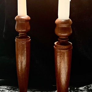 May include: Two wooden candle holders with a dark brown finish. Each holder has a round base and a tall, slender stem. The candles are white and are lit.