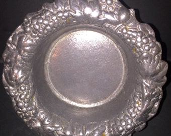 May include: A silver metal bowl with a grapevine design. The bowl is round and has a raised rim. The grapevine design is detailed and includes leaves and grapes.