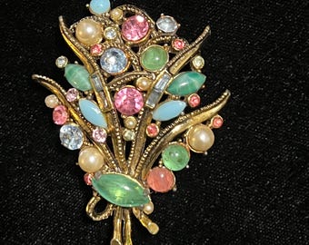 May include: A gold-toned brooch shaped like a floral bouquet. It is adorned with a variety of colourful gemstones, including pink, blue, green, and pearl-like beads. The brooch is set against a black backdrop.