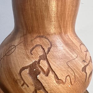 May include: A brown ceramic vase with a rounded body and a narrow neck. The vase features hand-painted brown figures and abstract designs. The figures appear to be engaged in a ritual or hunting scene. The vase is likely a decorative item.