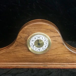May include: A wooden clock with a brass face and black hands. The clock is set on a wooden base with a curved top.