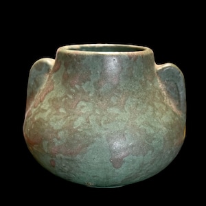 May include: A handcrafted, green ceramic vase with two side handles. The vase has a rounded body and a textured surface with brown and green color variations. The opening is circular, and the background is black.