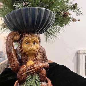 May include: A ceramic monkey figurine with a dark blue bowl on its head, holding a fruit. The monkey is brown and tan with a detailed face. The bowl holds a floral arrangement with green and yellow accents.
