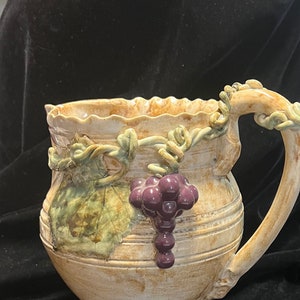 May include: A beige ceramic pitcher with a decorative handle and a cluster of purple grapes. The pitcher has a textured surface and is adorned with green leaves and vines. The rim has a scalloped edge.