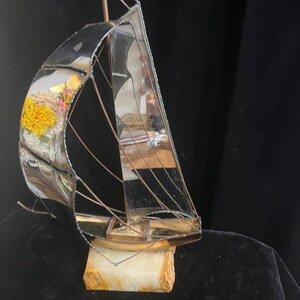 May include: A decorative sailboat sculpture with a mirrored finish, copper-colored rigging, and a small flag. The boat is mounted on a rectangular, cream-colored stone base. The sails reflect the surrounding environment.