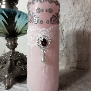 May include: A pink pillar candle with white lace and silver glitter accents. The candle has a red jewel and a pearl hanging from it.