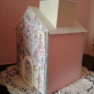 Op de afbeelding: Een roze en witte huisvormige doos met een bloemenpatroon en een witte schoorsteen. De doos heeft een zilveren rand en een kleine witte deur.