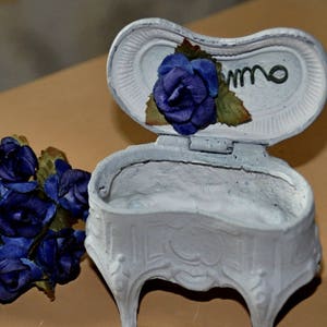 May include: A white ceramic trinket box with a hinged lid. The lid is decorated with a blue fabric flower and green leaves. The box has a decorative, ornate base with four legs. The box is sitting on a brown surface.