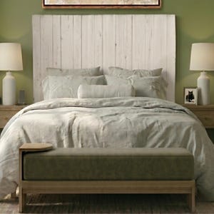 May include: A rectangular upholstered bench with a muted olive green seat and a light wood frame. The bench features a small, integrated circular side table on the left end. The piece is positioned at the foot of a bed with a white headboard.